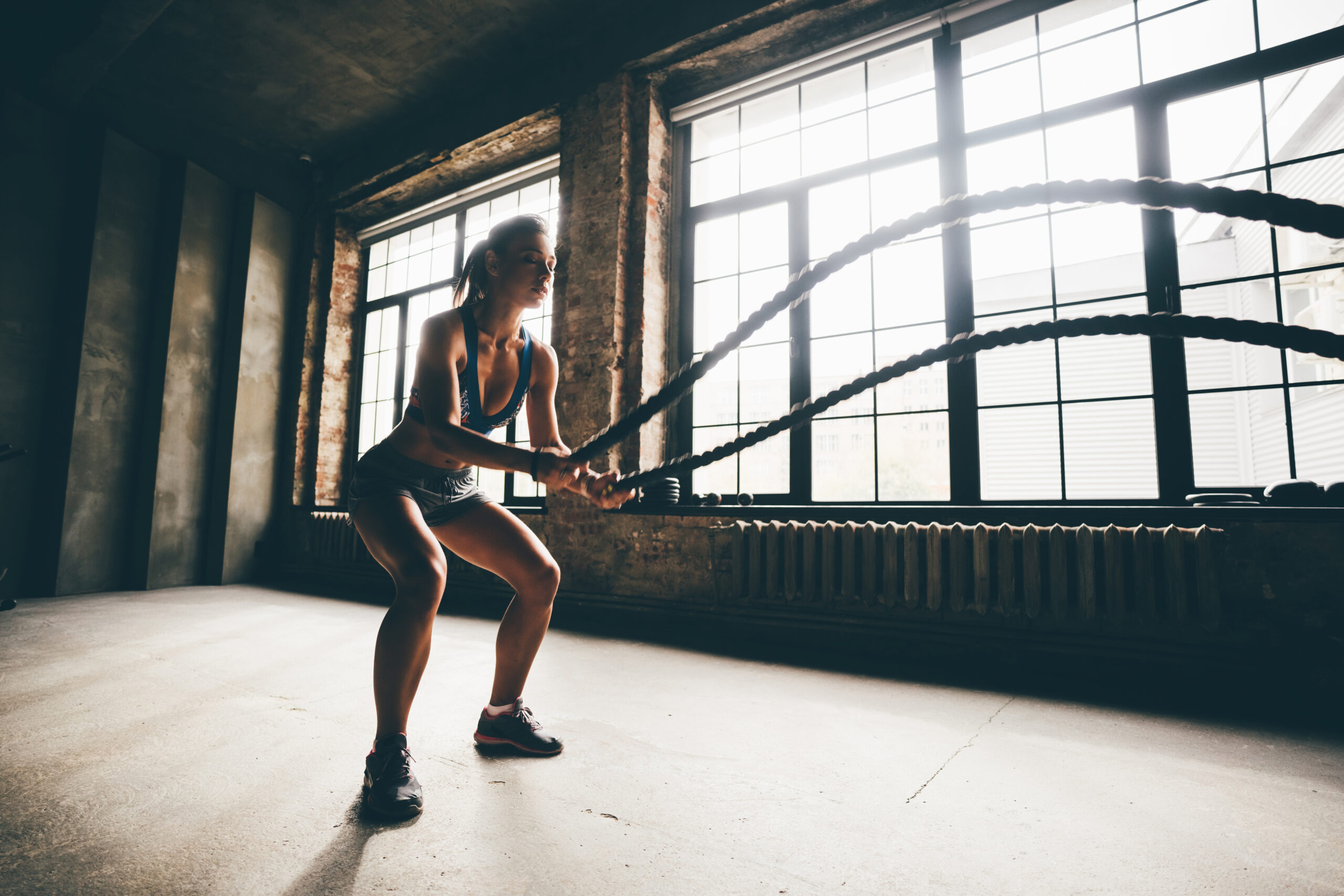 Fitness girl exercising with battle ropes at gym. Woman training doing battling rope workout working out arms and cardio for cross fit exercises.